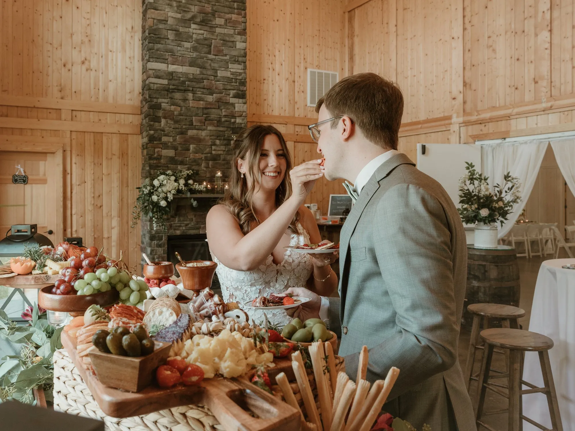 Newlyweds sharing a charcuterie spread by the stone fireplace in the cocktail room at The Heritage Barn