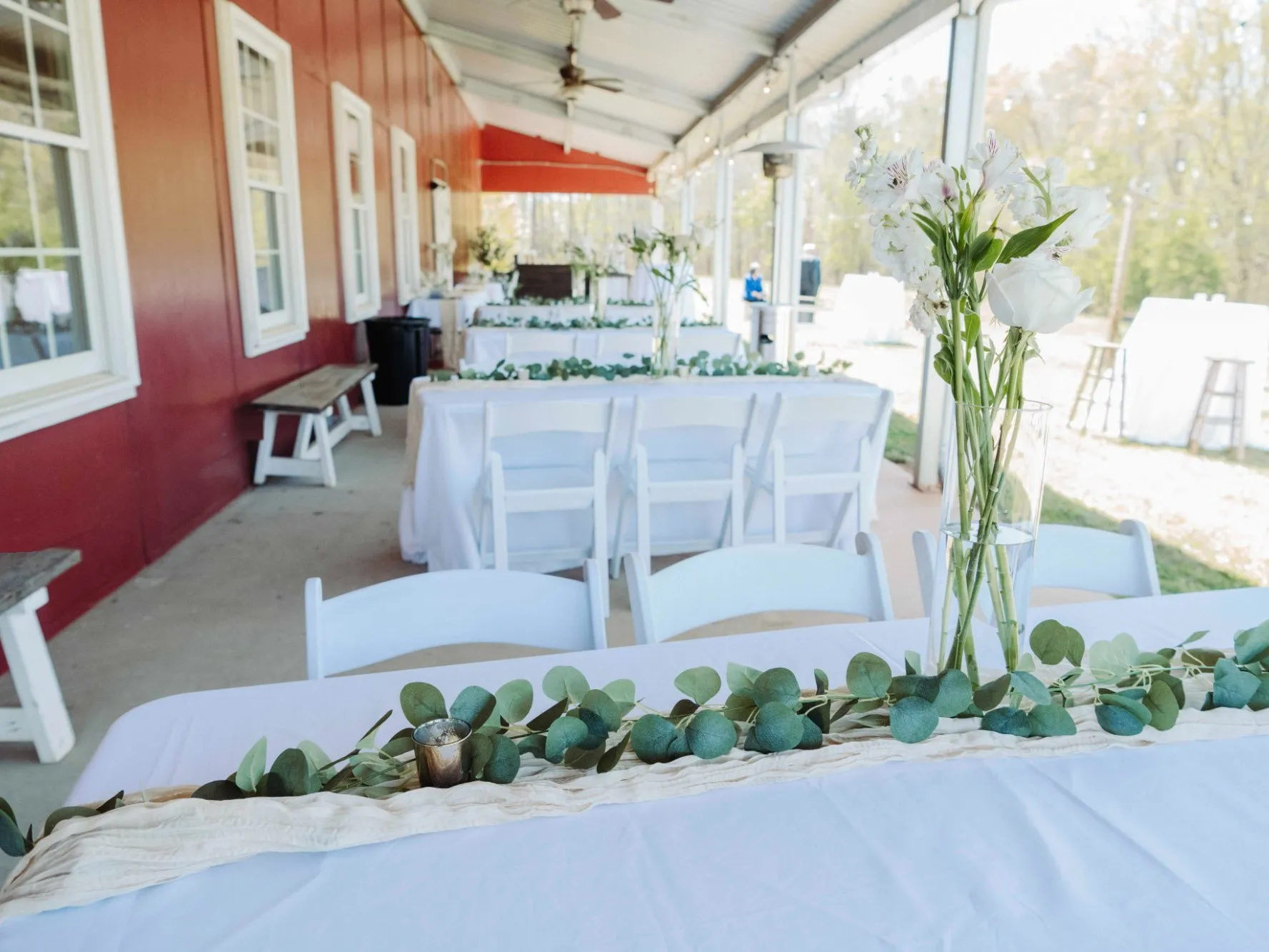 Covered patio set for a reception with white linens, eucalyptus garland and fresh flowers along the red barn at The Heritage Barn