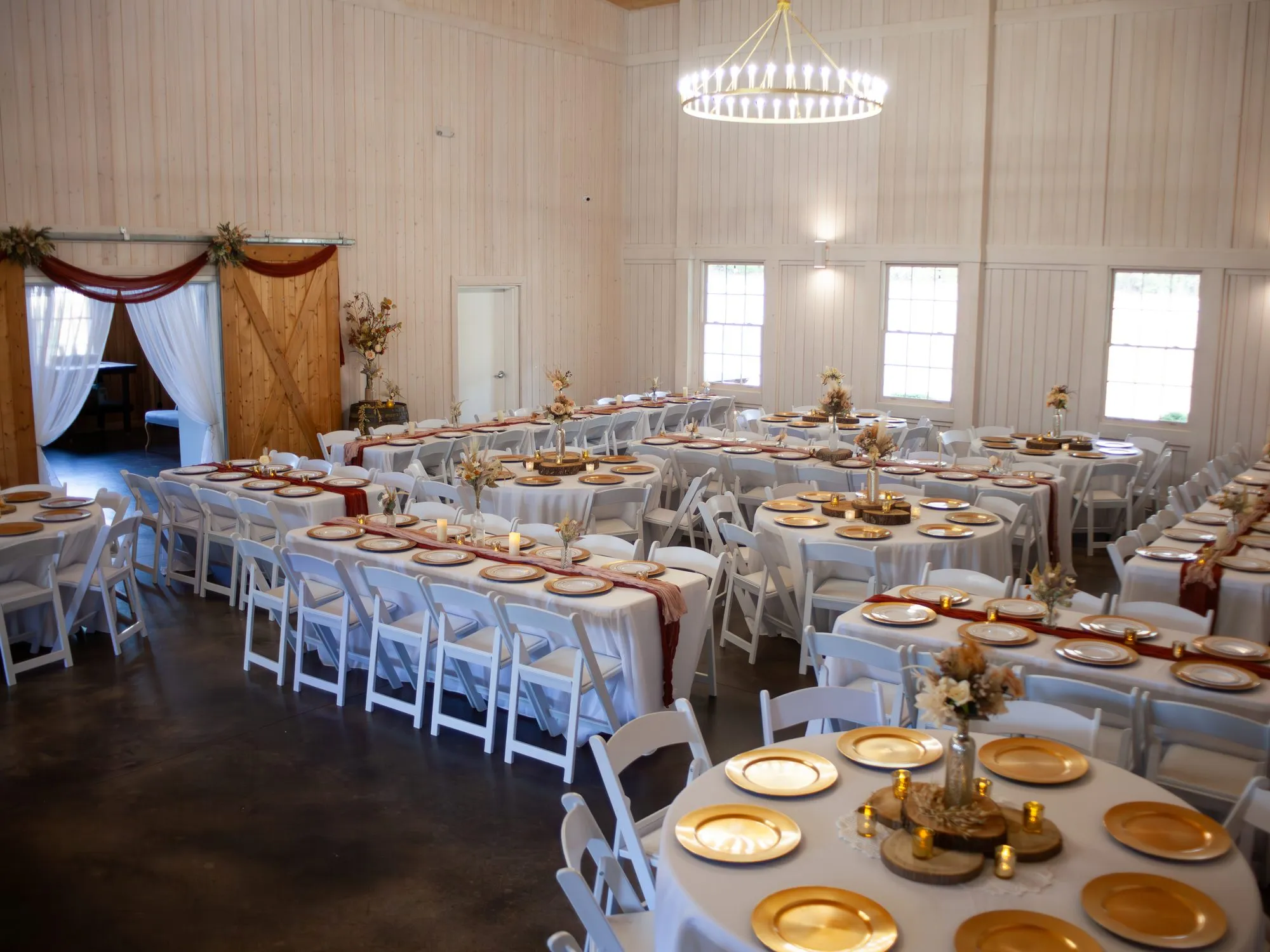 Barn reception set with white linens and gold chargers beneath the chandelier at The Heritage Barn