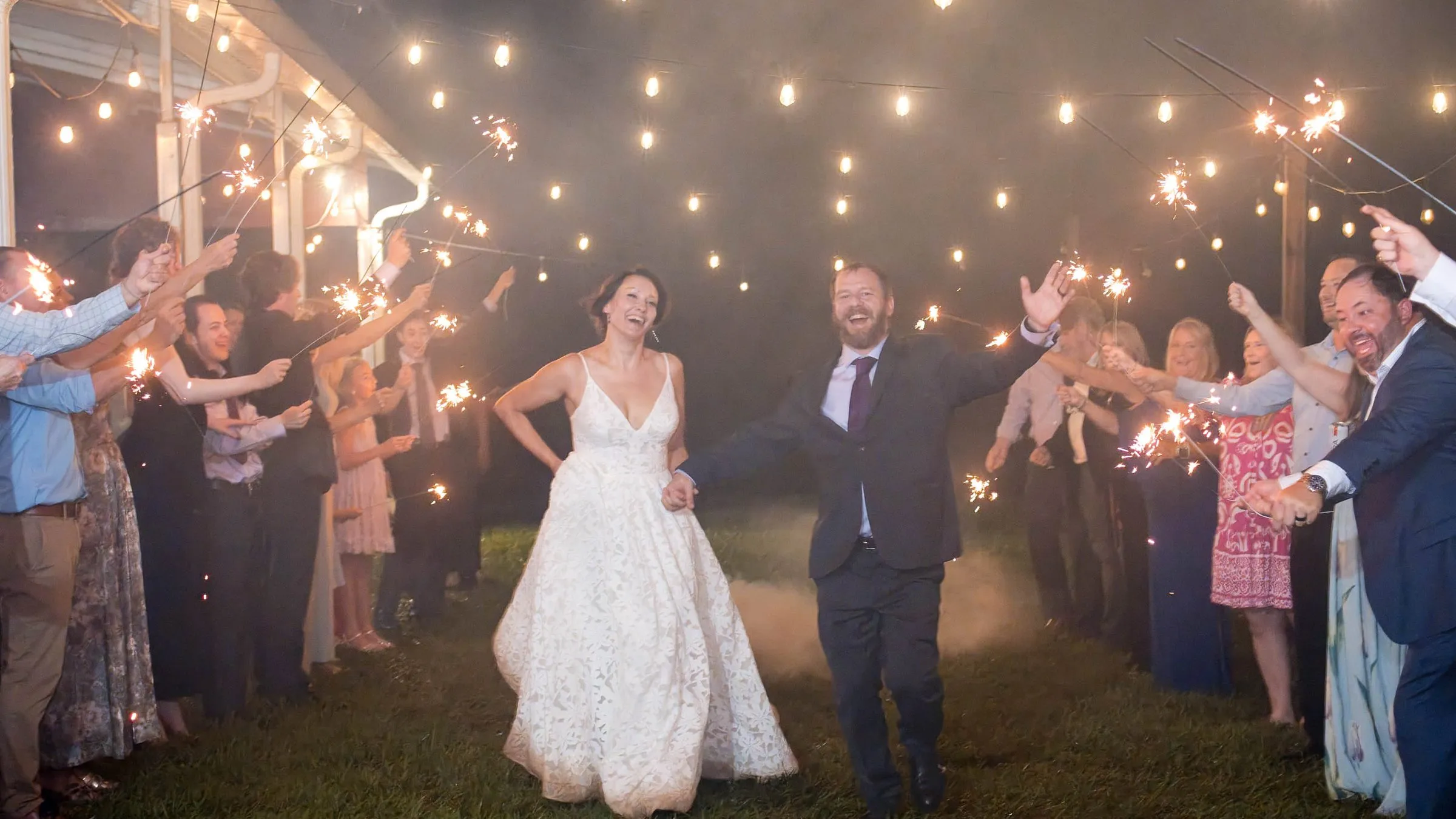 Newlyweds running through a sparkler send-off under the patio string lights at The Heritage Barn